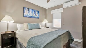 Une chambre à coucher avec un lit, deux lampes de chevet, une table de nuit, une fenêtre avec des stores et un tableau représentant des voiliers accroché au mur.