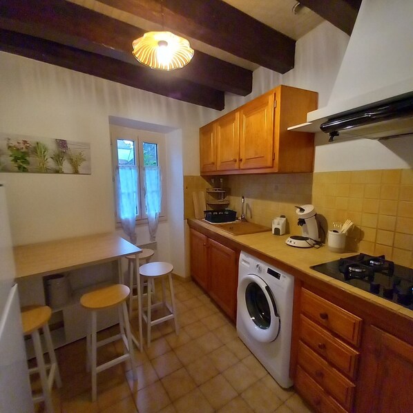 A compact kitchen with wooden cabinets, a washing machine, a sink, and a small dining area with stools.