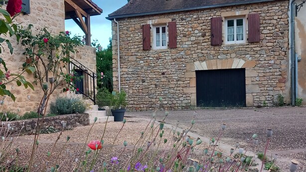 A stone building with a garage door, surrounded by a gravel driveway and various flowering plants.