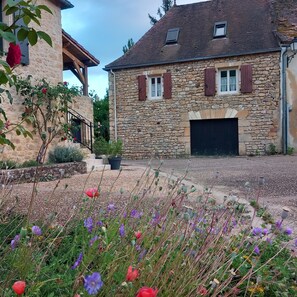 A stone building with a garage door, surrounded by a gravel driveway and various flowering plants. - Le logis d'Epicure Charming house in the heart of a typical Lot village. (Payrignac)