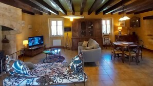 A spacious living room with a sofa, a coffee table, and a television. There are wooden beams on the ceiling and a dining area with chairs and a table.