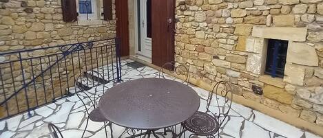 Un bâtiment en pierre doté d’une terrasse carrelée, d’une table ronde entourée de chaises et d’une petite fenêtre avec des volets.