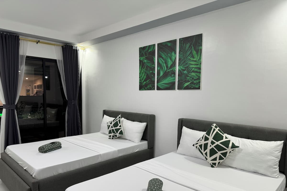 A hotel room with two single beds, a large window with curtains, and a wall with three green leaf-patterned paintings.