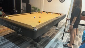 Een pooltafel met ballen, een persoon staat ernaast en een dartbord hangt aan de muur.