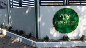 Een bord voor Steffen Resort met een rond logo met palmbomen en een huis, omgeven door een witte muur en kleine planten.