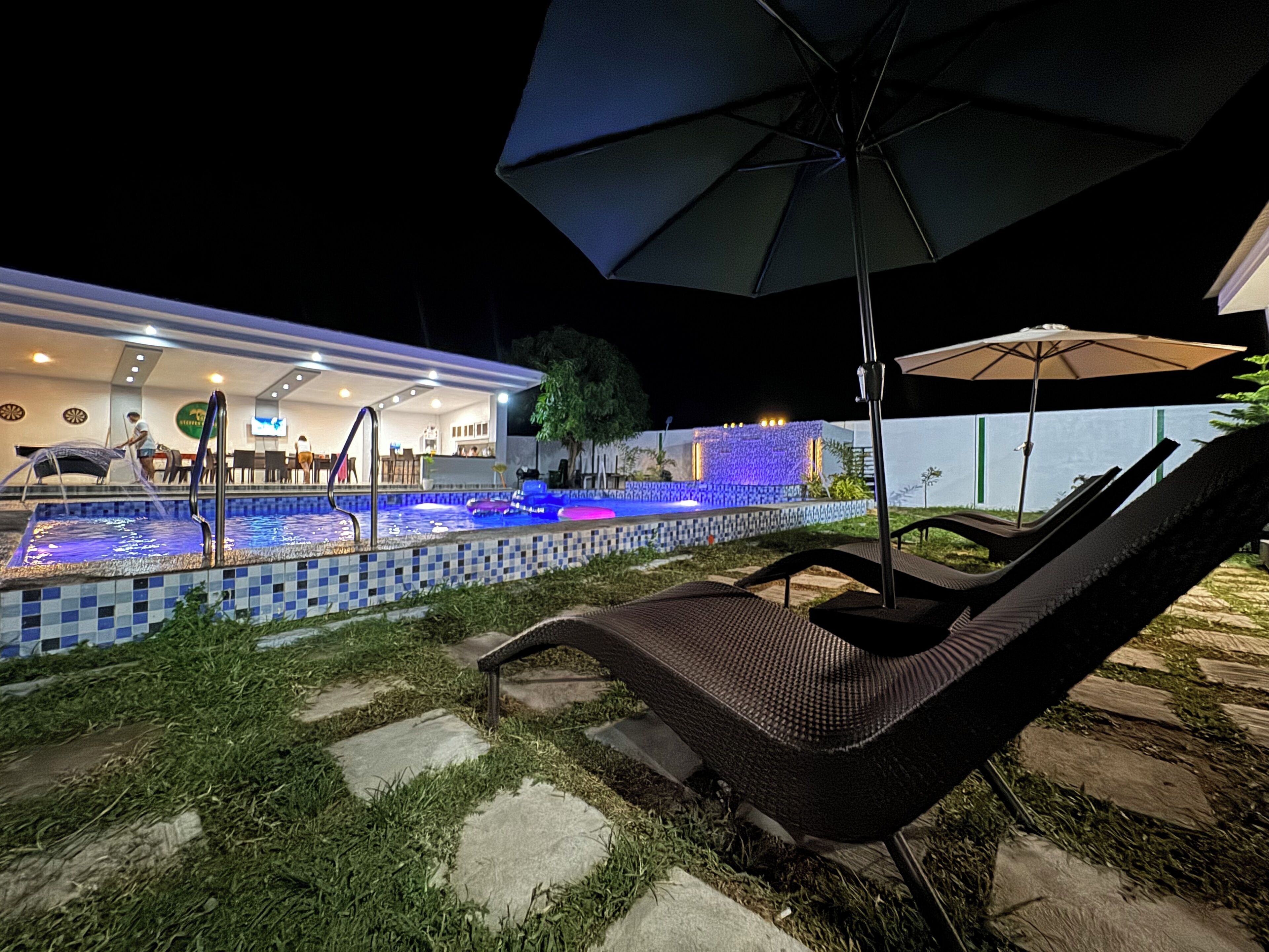 Zona de piscina amb gandules i ombrelles, solàrium amb rajoles, i un edifici al fons amb bar i zona d'estar.