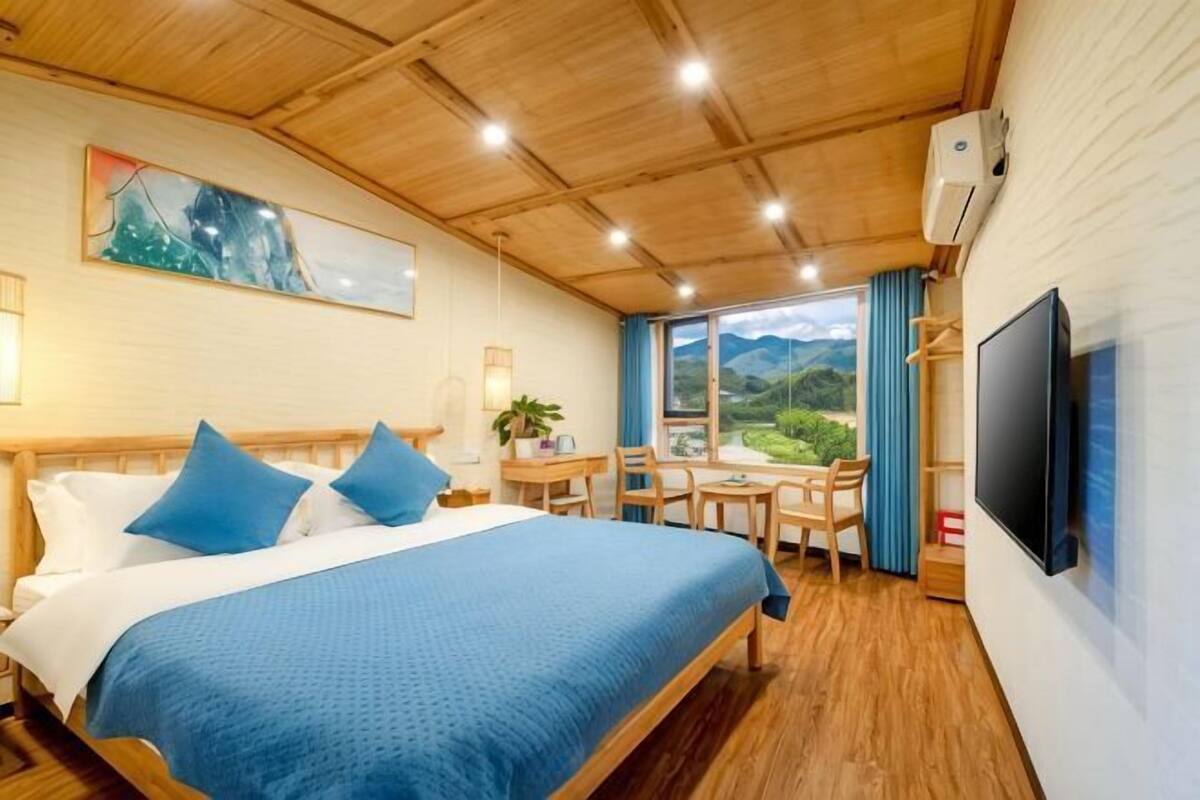 A hotel room with a bed, a desk, a chair, a TV, and a window with a view of greenery.