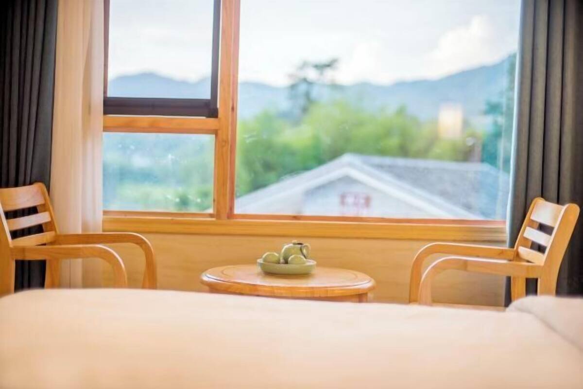 A room with a bed, two wooden chairs, a small round table, and a view of mountains through a large window.