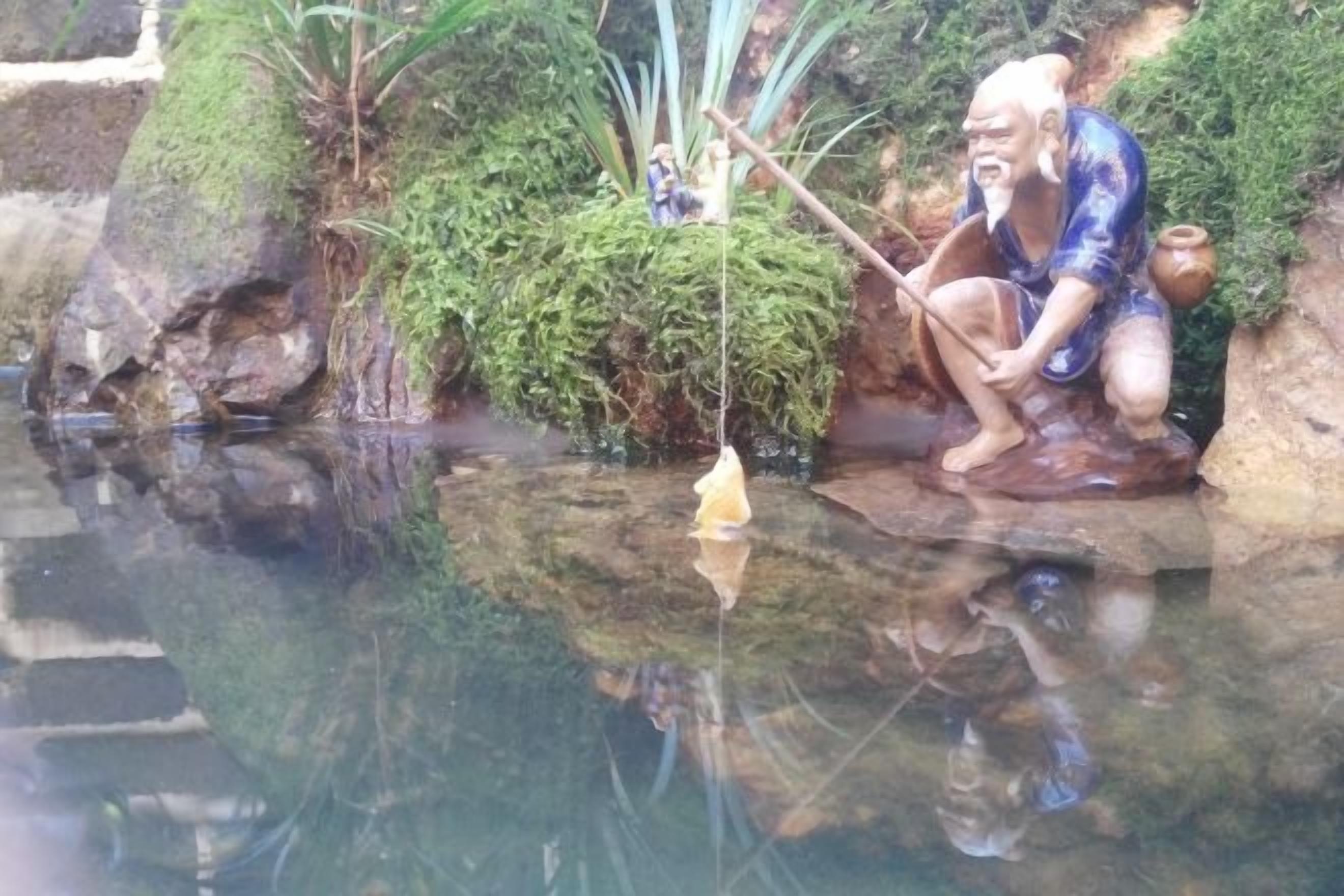 A serene pond with a statue of a man fishing, surrounded by lush greenery and moss-covered rocks.