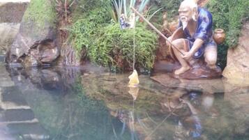 A serene pond with a statue of a man fishing, surrounded by lush greenery and moss-covered rocks.