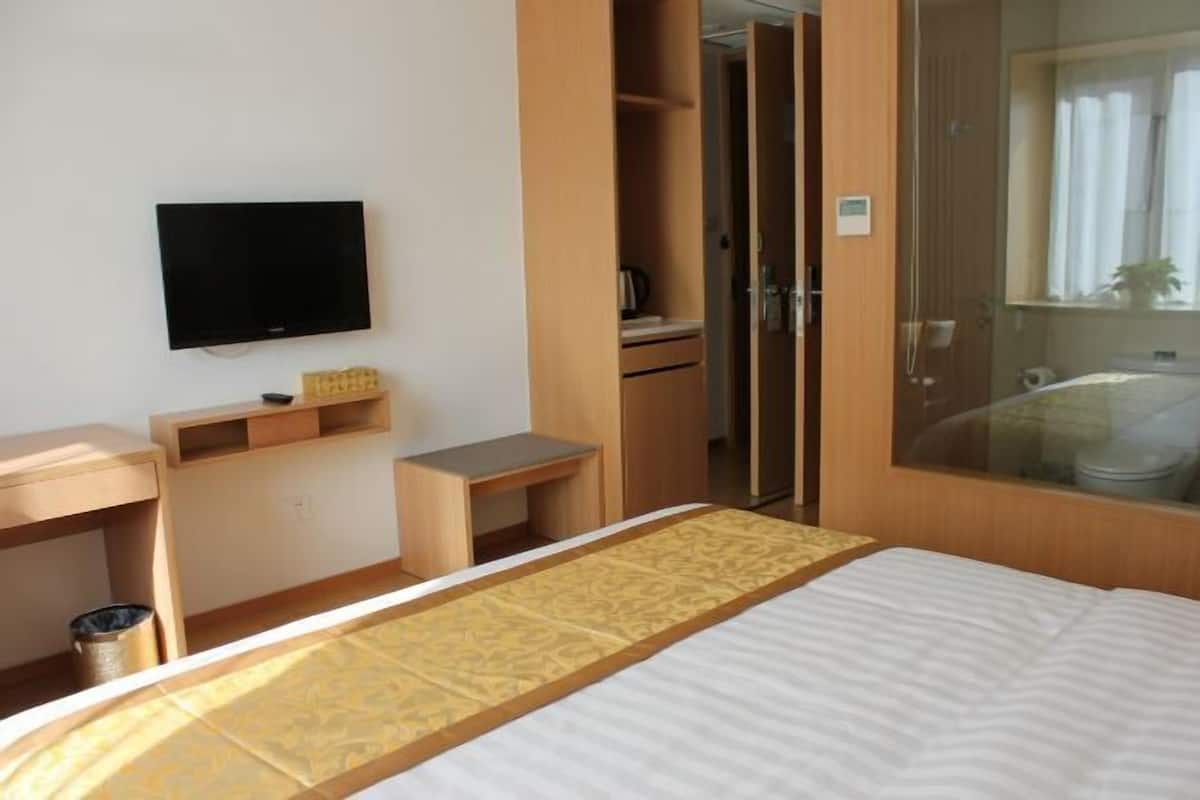 A hotel room with a bed, a television, a desk, a wardrobe, and a bathroom visible through a glass partition.