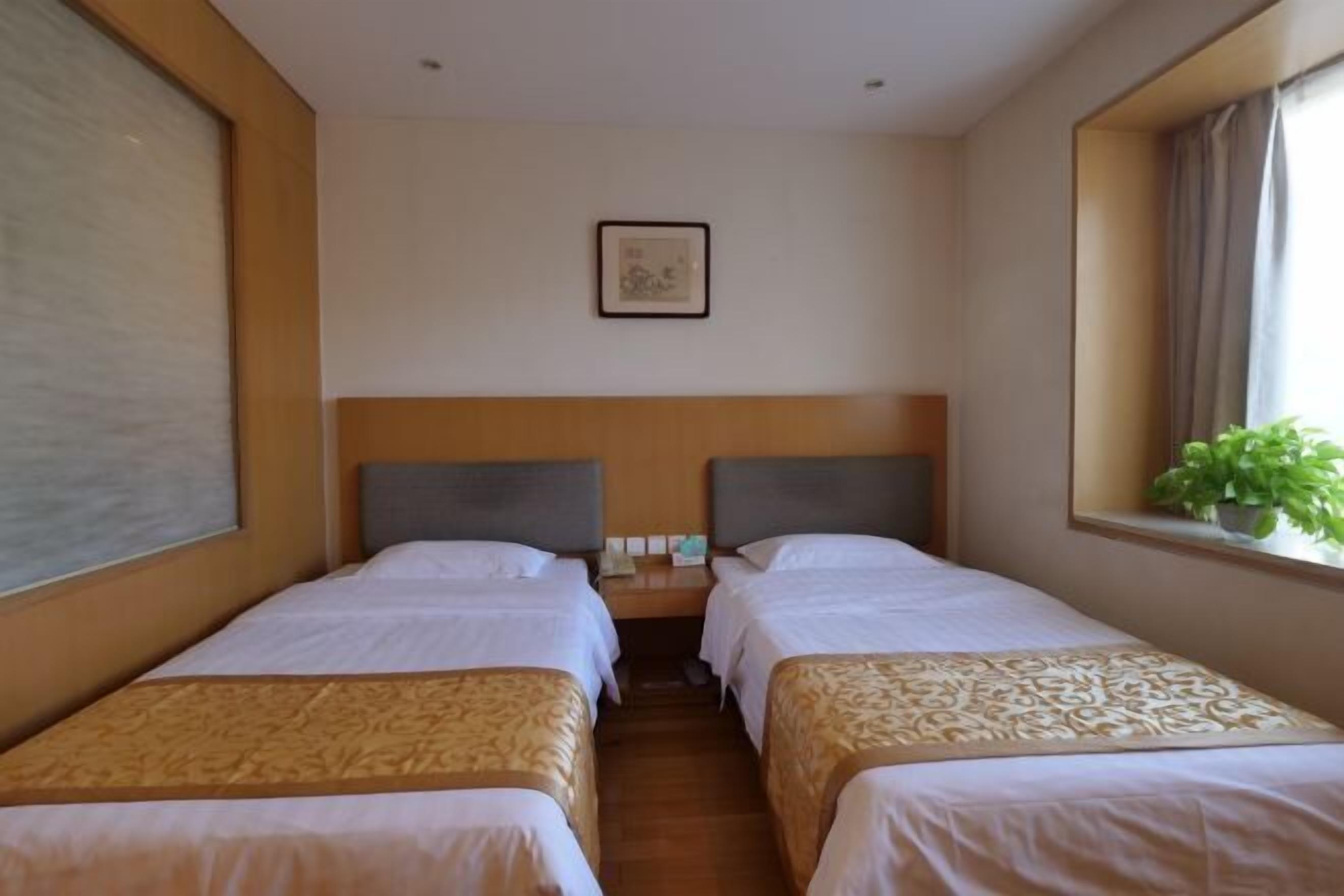 A hotel room with two single beds, a wooden headboard, a small bedside table, a framed picture on the wall, and a window with a plant.