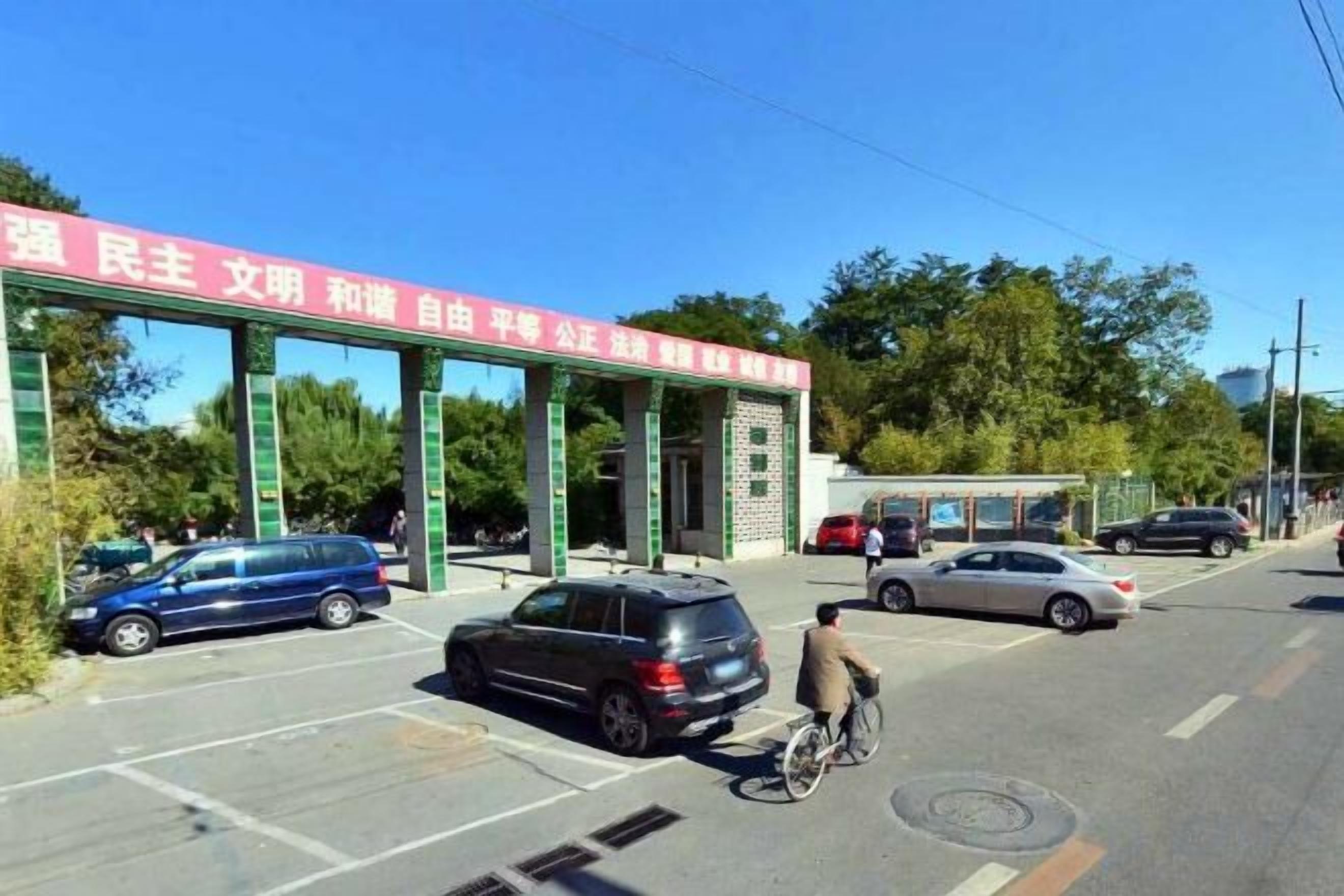 Un edificio con un estandarte rojo, un estacionamiento con coches y una persona montando en bicicleta.