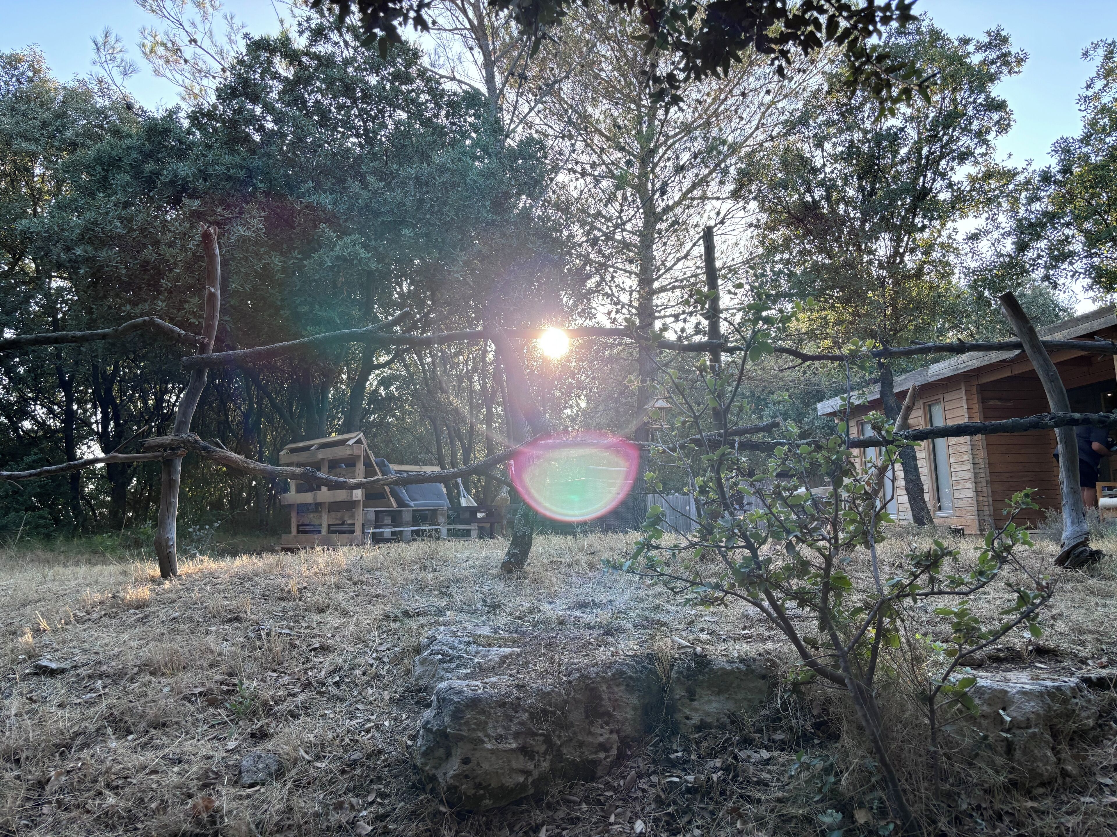 A rustic cabin with a wooden deck, surrounded by trees and a stone wall.