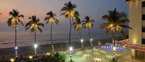 Un resort con piscina, zona de estar al aire libre, palmeras y vistas a la playa al atardecer.