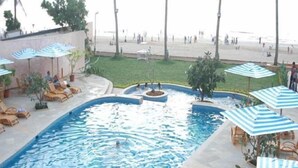 A swimming pool with lounge chairs and umbrellas, a beach in the background, and palm trees.