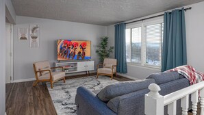 A living room with a television, two armchairs, a sofa, a white console table, and a large window with curtains. - NEW! Charming Duplex - Central, Ski, Hike, Unwind! (Sandy)