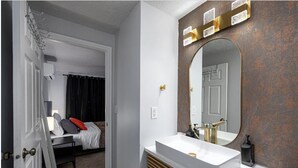 A hotel room with a bed, a bedside table, a mirror, and a bathroom with a sink and mirror. - NEW! Charming Duplex - Central, Ski, Hike, Unwind! (Sandy)