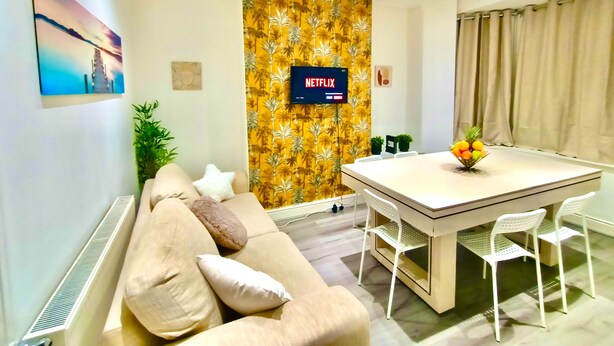 Ett modernt vardagsrum med en soffa, ett matbord med stolar, en tv som är monterad på en vägg med blommig tapet och en växt.
