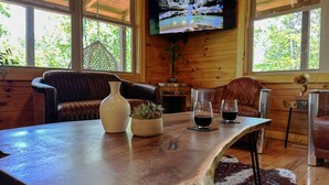 Ein gemütliches Blockhaus-Interieur mit einem Holztisch, Ledersesseln, einem an der Wand montierten Fernseher und einem großen Fenster mit Blick auf Bäume.