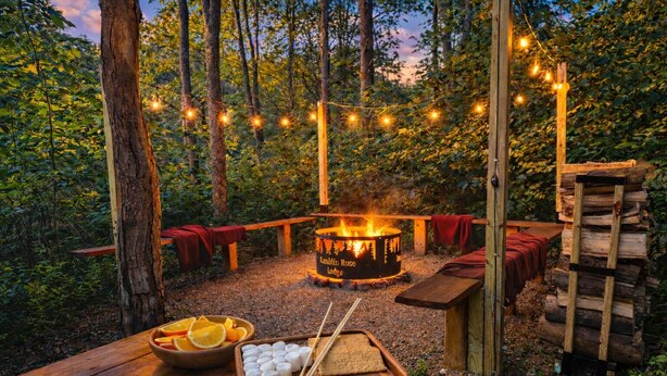 Ein Lagerfeuer mit einem Schild, einer hölzernen Bank, einem Tisch mit Snacks und Lichterketten im Wald.