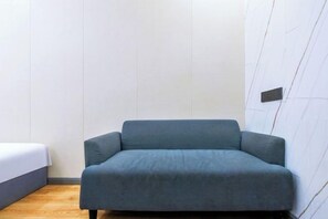 A room with a single grey sofa, a bed with white bedding, and a wooden floor.