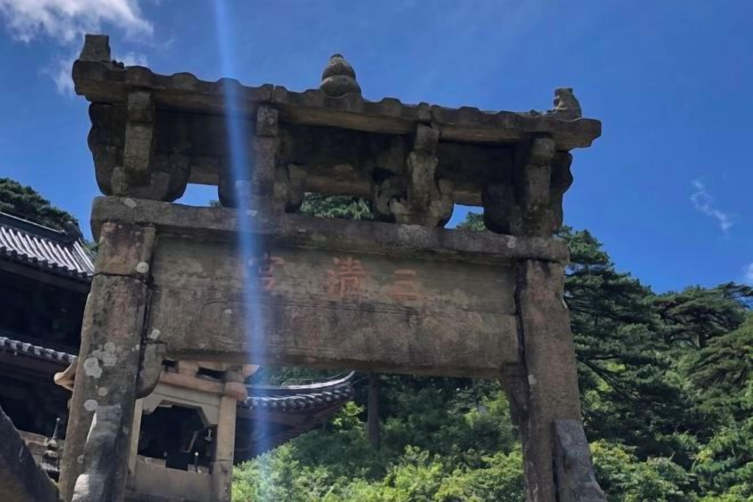 A traditional stone structure with inscriptions, possibly a historical monument or gate, set against a backdrop of lush greenery and a clear sky.