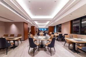 En hotellrestaurang med flera bord och stolar, ett stort, runt bord i mitten och utsikt över stadsbilden genom stora fönster.