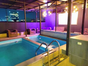 An indoor swimming pool with a view of a cityscape at night, surrounded by purple walls and seating areas. - $ PROMO $ 11 Bedrooms with bathrooms Aprtmnt + 2 Jacuzzis and a rooftop pool. @Poblado (Medellín)
