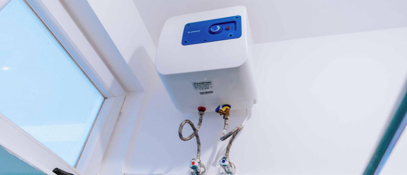 A white air conditioning unit with a blue control panel, two water valves, and a window to the left.