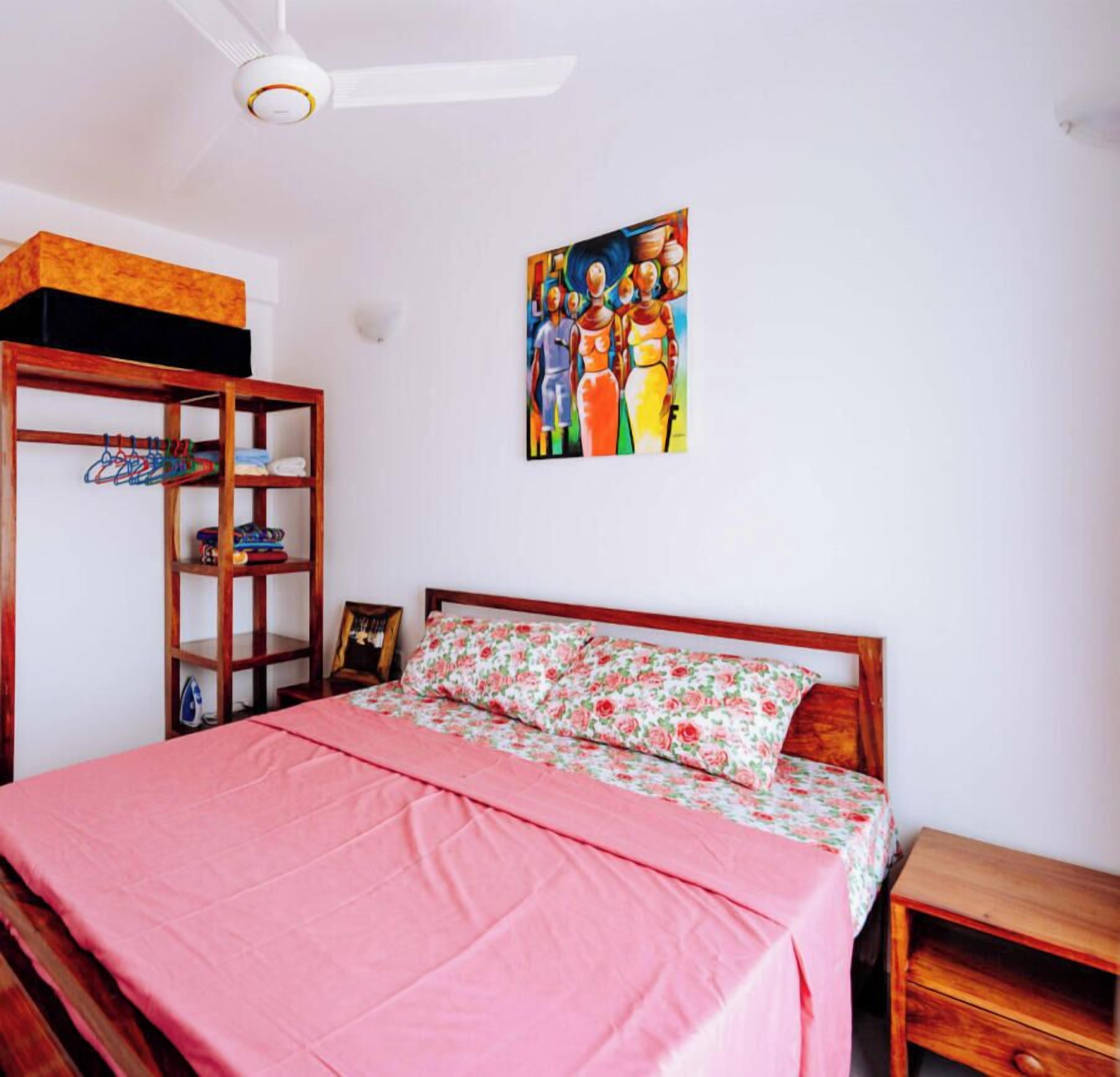 A bedroom with a bed, a wooden nightstand, a shelf with hanging clothes, and a ceiling fan.