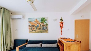 A living room with a blue sofa, a wooden coffee table, a painting of a market scene, and a ceiling fan. - Beautiful ocean-view 2 bedroom home in Fumba Town (Fumba Town)