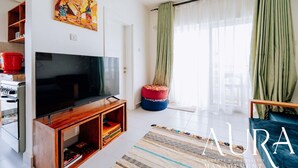 A modern living room with a flat-screen TV on a wooden stand, a colorful painting on the wall, a green curtain, and a patterned rug. - Beautiful ocean-view 2 bedroom home in Fumba Town (Fumba Town)