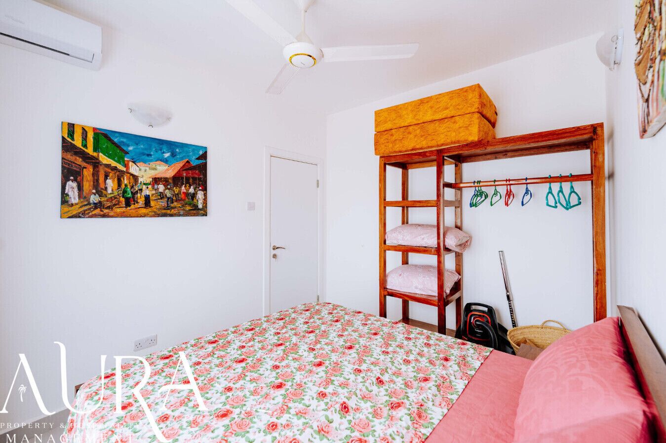 A bedroom with a floral bedspread, a wooden shelf with hanging items, and a painting on the wall.