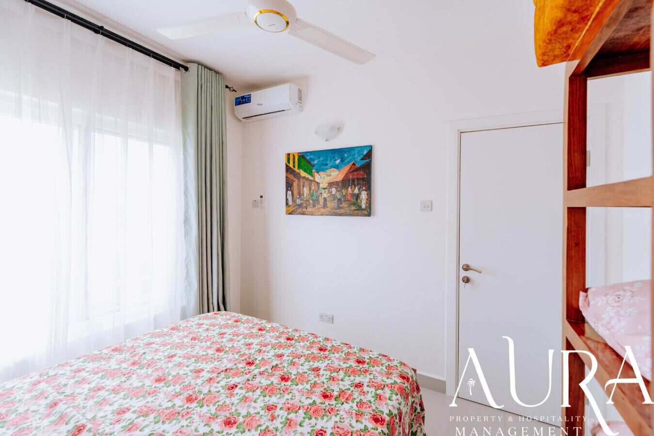A bedroom with a bed, a ceiling fan, an air conditioning unit, a door, and a painting on the wall.