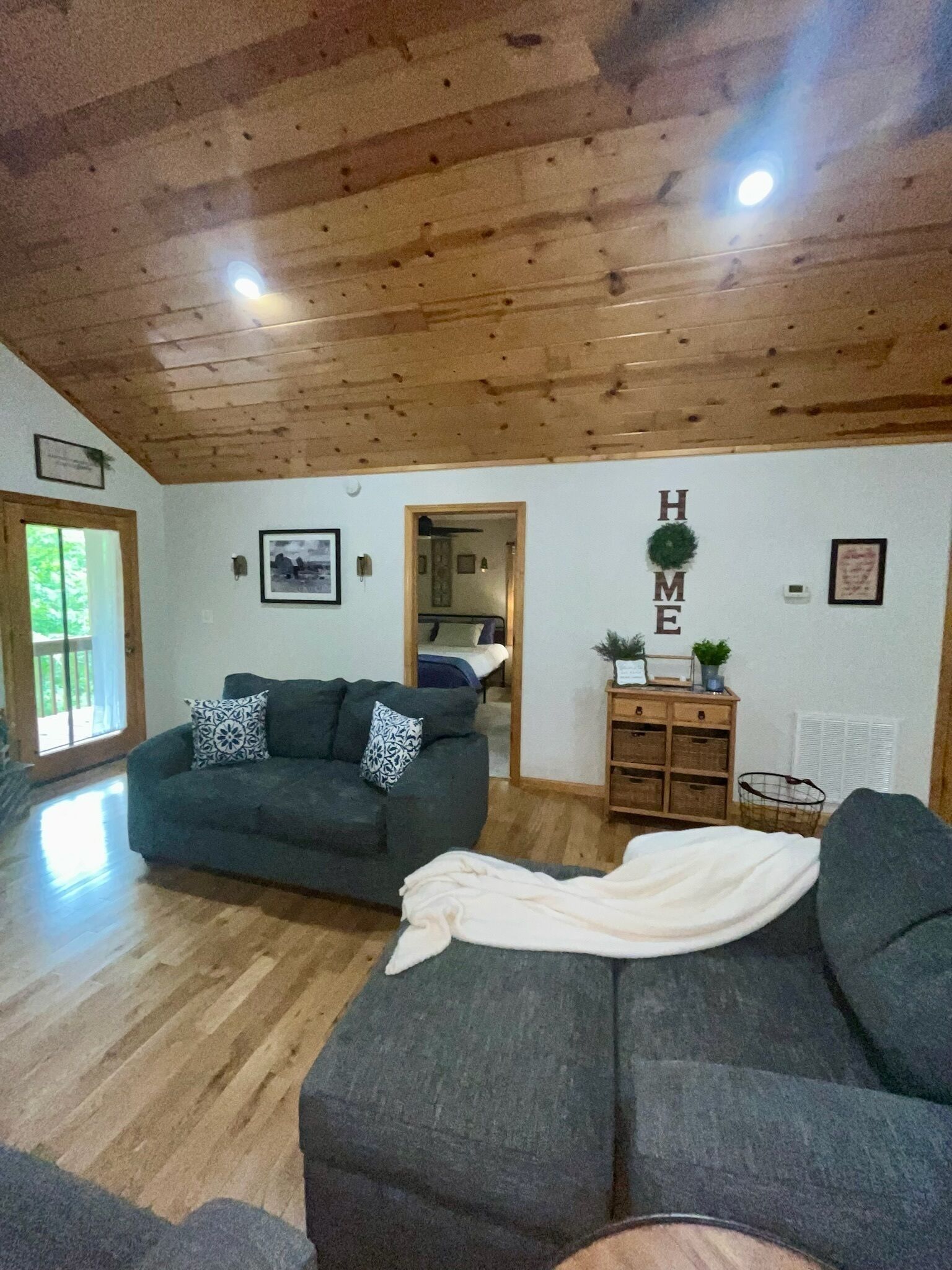 Un salon avec un plafond en bois, un canapé gris, un fauteuil blanc et un petit meuble en bois.