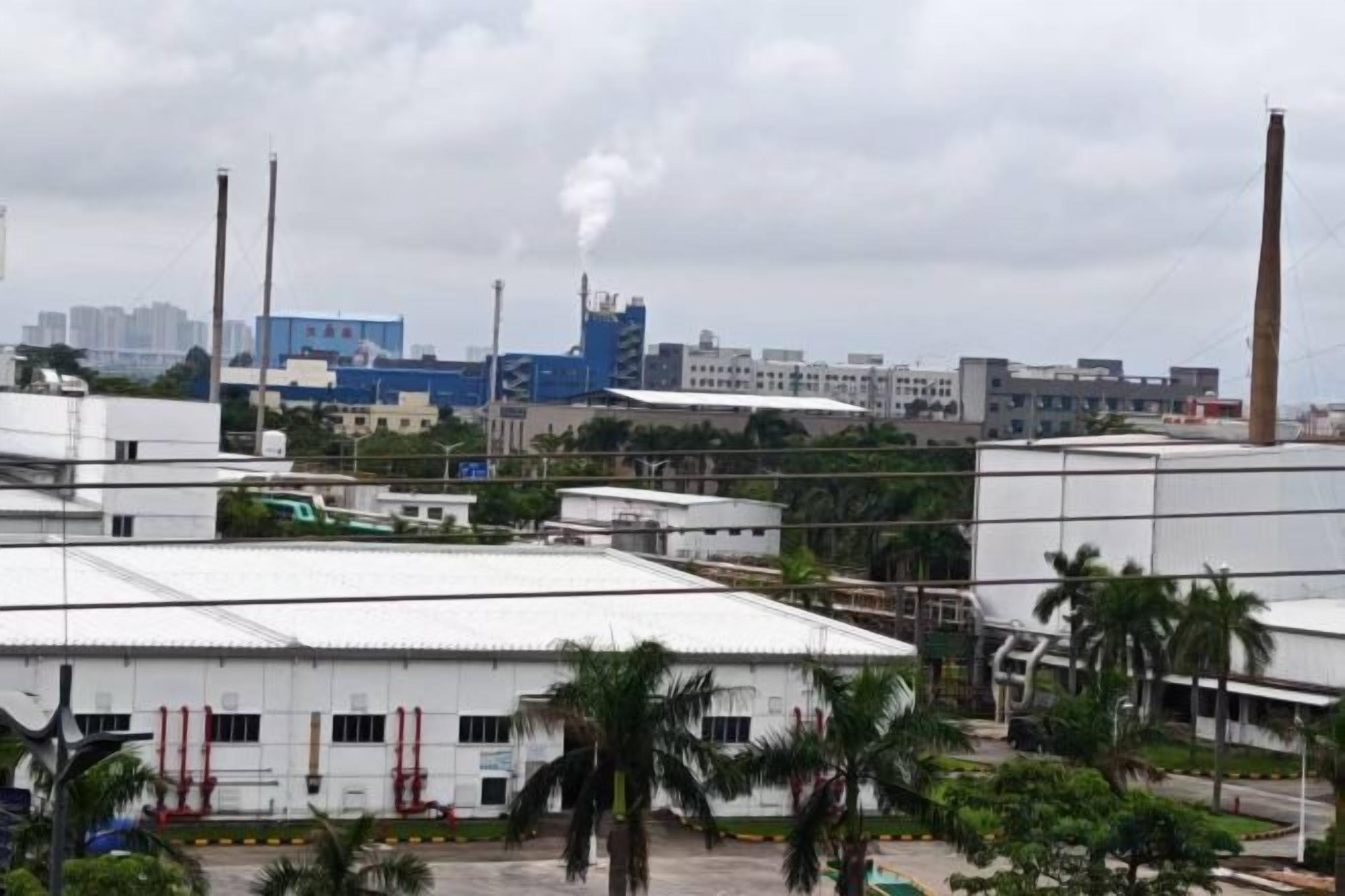 Uma extensa área industrial com vários edifícios, incluindo uma chaminé alta e imponente expelindo fumaça.