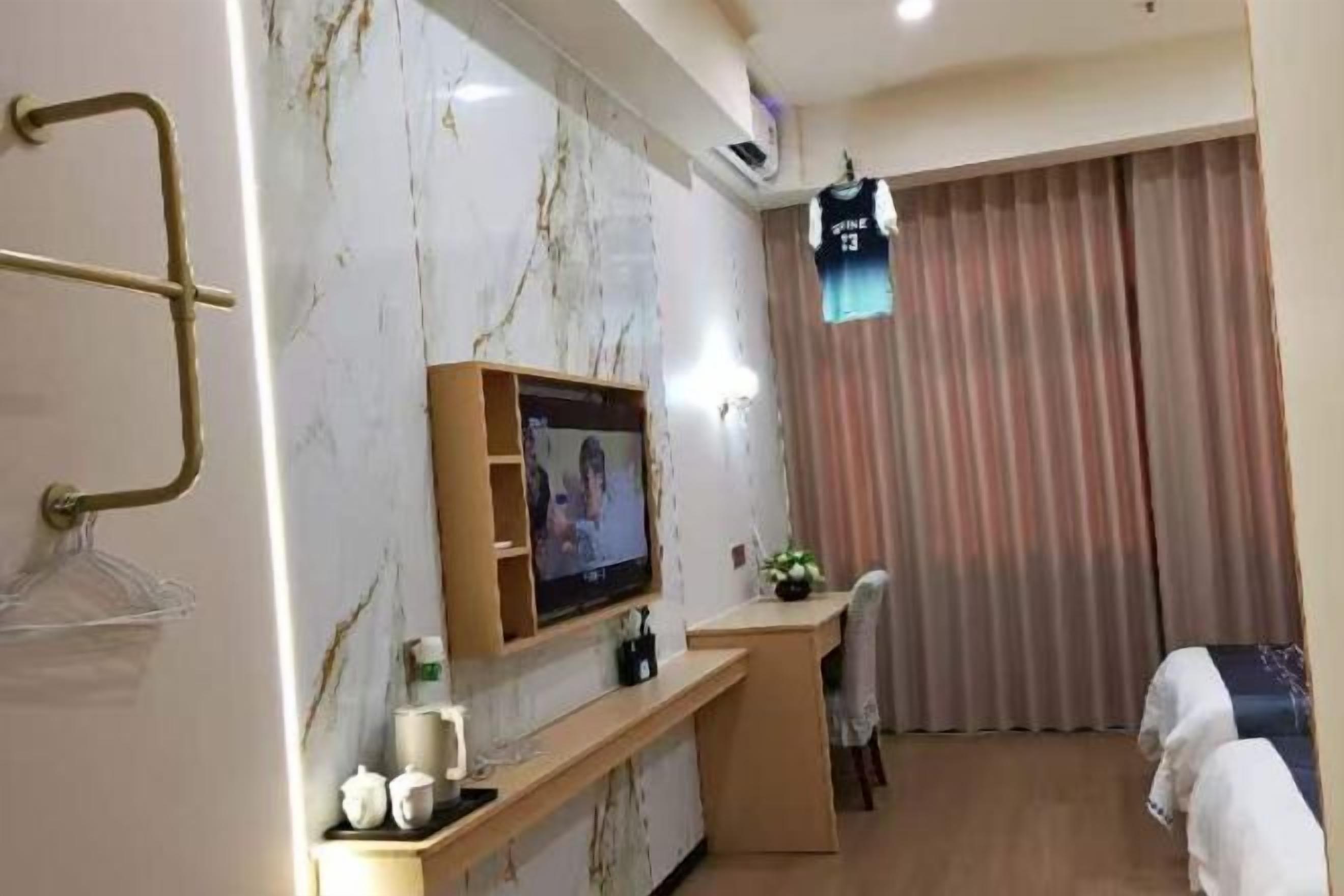 A hotel room with a TV mounted on the wall, a desk with a chair, a bed with towels, and a window with curtains.