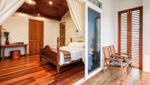A bedroom with a wooden bed, a nightstand, a chair, and a sliding glass door leading to a balcony. - Chiang Mai Colonial House (Tambon San Phi Suea)