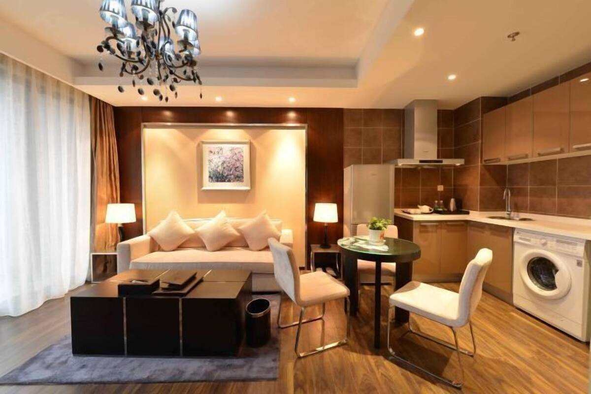 A modern living room with a sofa, coffee table, and chairs, a dining table with a lamp, and a kitchen area with a washing machine.