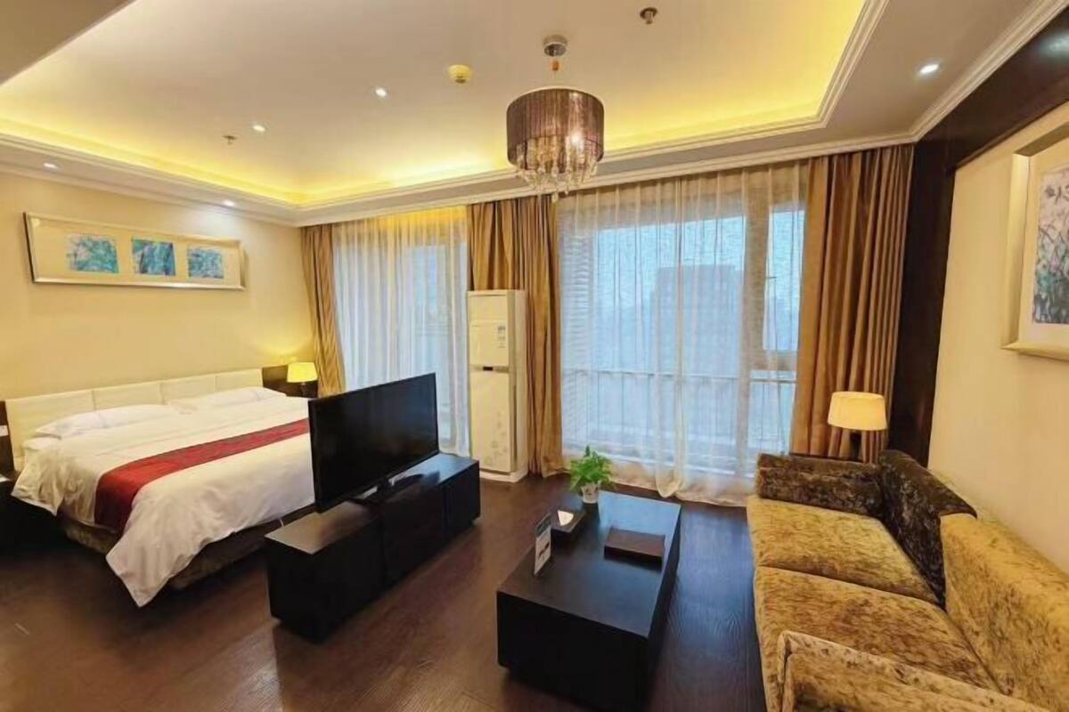 A hotel room with a bed, a TV, a sofa, a coffee table, and a large window with curtains.