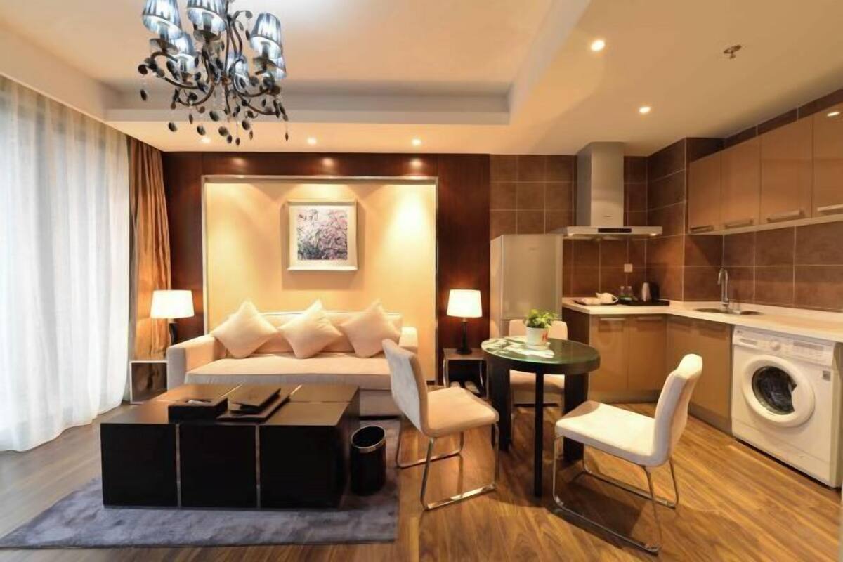 A modern living room with a sofa, coffee table, and chairs, a dining table, a chandelier, and a kitchen area with a washing machine.