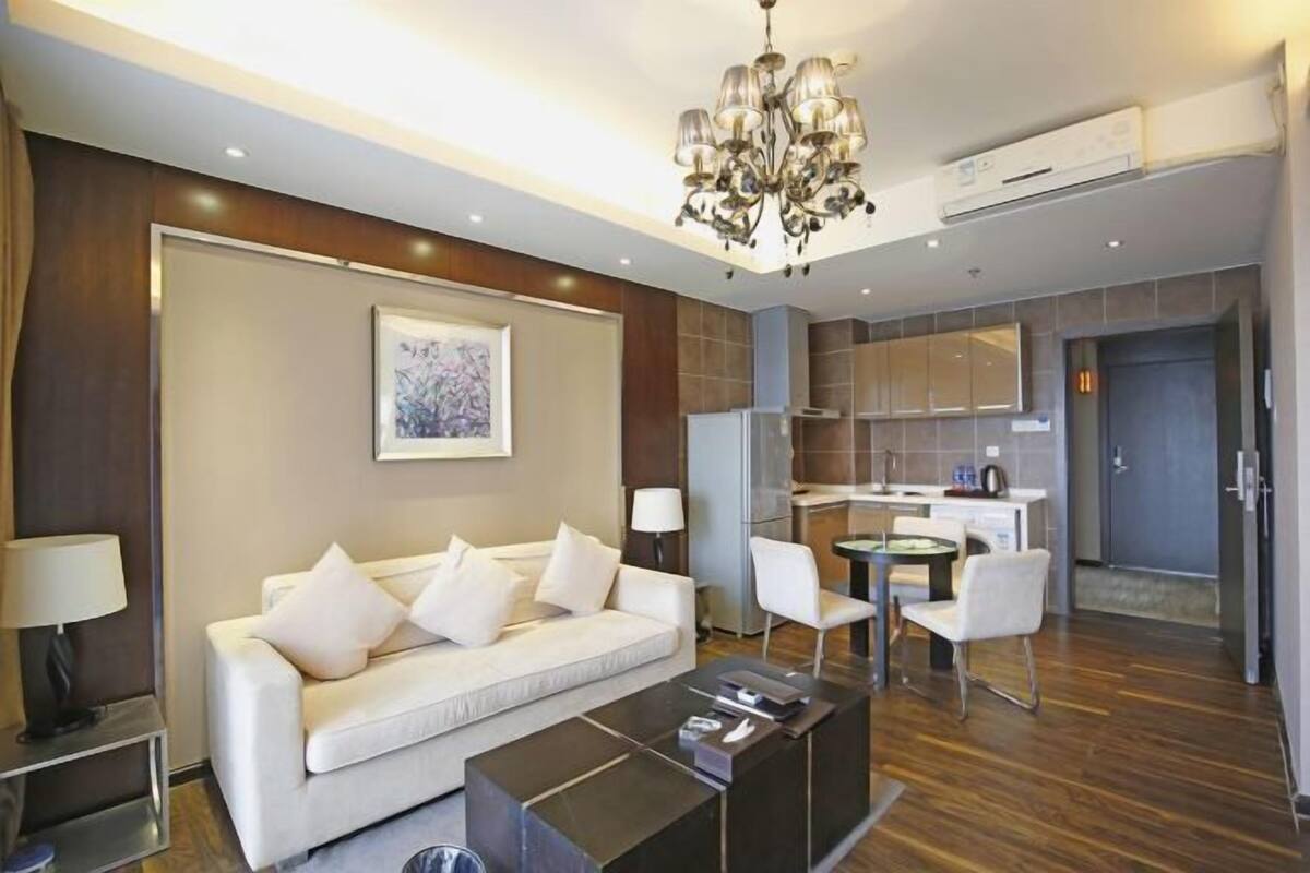 A modern living room with a sofa, coffee table, and a dining area with chairs. There is a kitchen area with cabinets and a refrigerator.