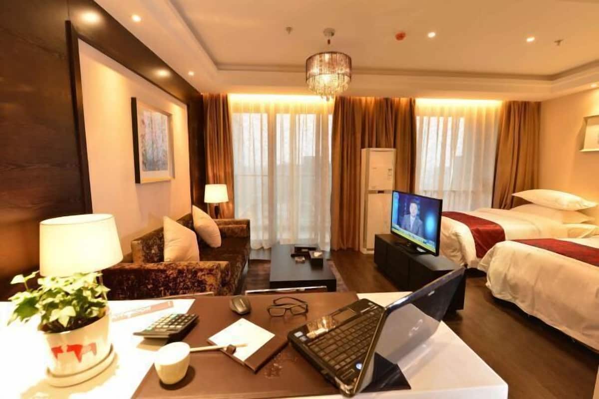A hotel room with a bed, a desk with a laptop, a TV, a sofa, a coffee table, and a potted plant.