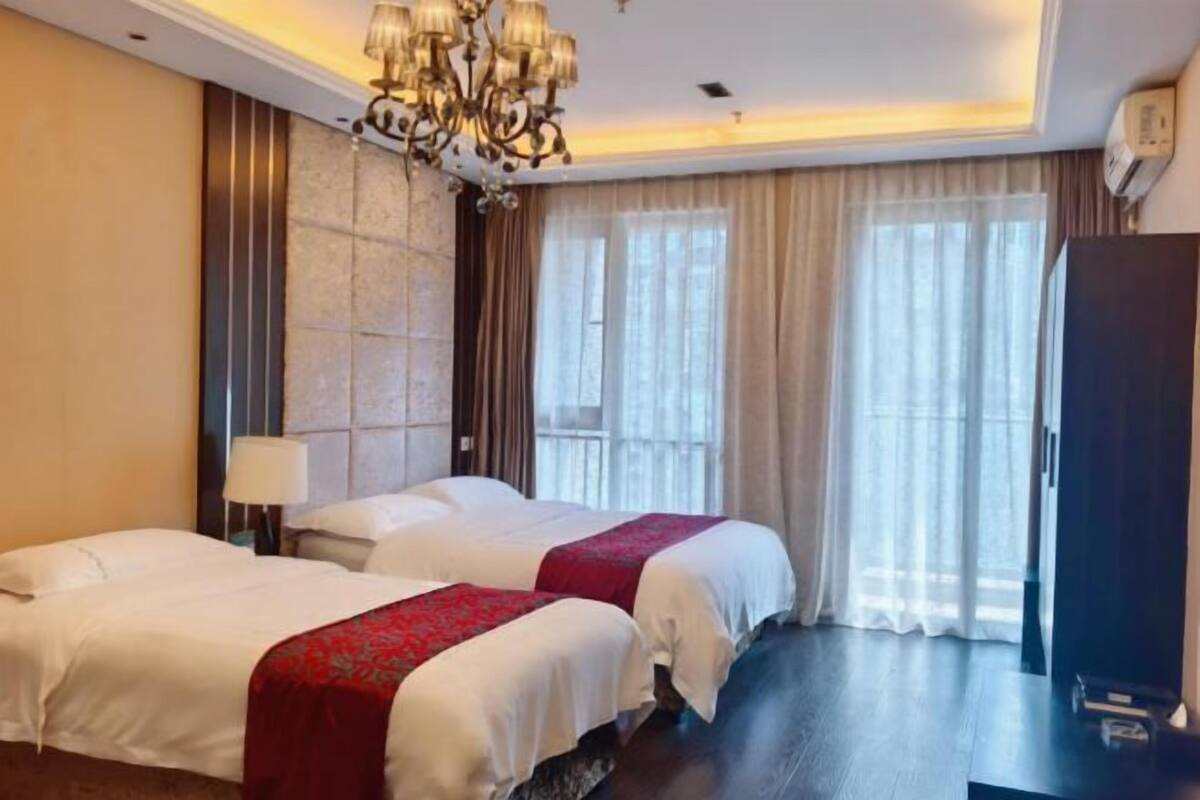 A hotel room with two beds, a chandelier, a large window with curtains, and a wardrobe.