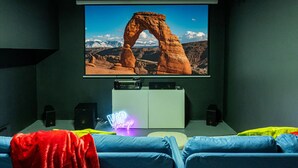 Cine en casa con una pantalla grande mostrando una escena de montaña, un mueble blanco con un letrero de neón y asientos azules.