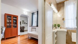Une pièce avec un plancher en bois, un canapé blanc, un meuble avec des portes en verre et une salle de bain équipée d’un lavabo et de murs carrelés.