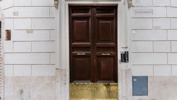 Une porte en bois à deux battants ornée d’un heurtoir en laiton, un mur blanc avec une plaque numérotée et une petite boîte électrique bleue.