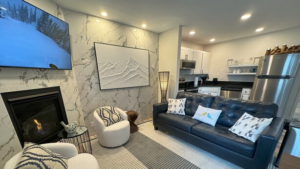 A modern living room with a fireplace, a sofa with decorative pillows, a mounted TV, and a kitchen area in the background. - Mountain Bluebird Condo Getaway at Schweitzer Ski Resort (Sandpoint)