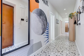 Un couloir avec une grande fresque murale, une porte en bois, un mur blanc avec un interrupteur et un sol carrelé.